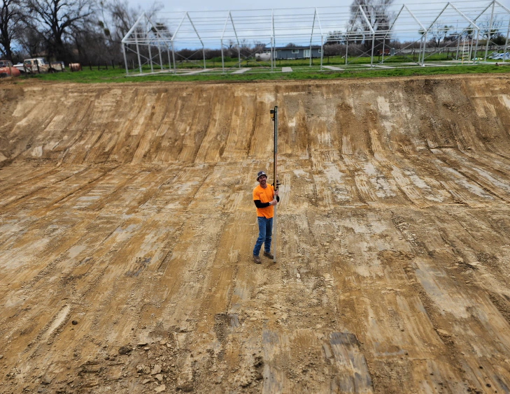 man wearing orange shirt in middle of excavated land and holding measuring equipment